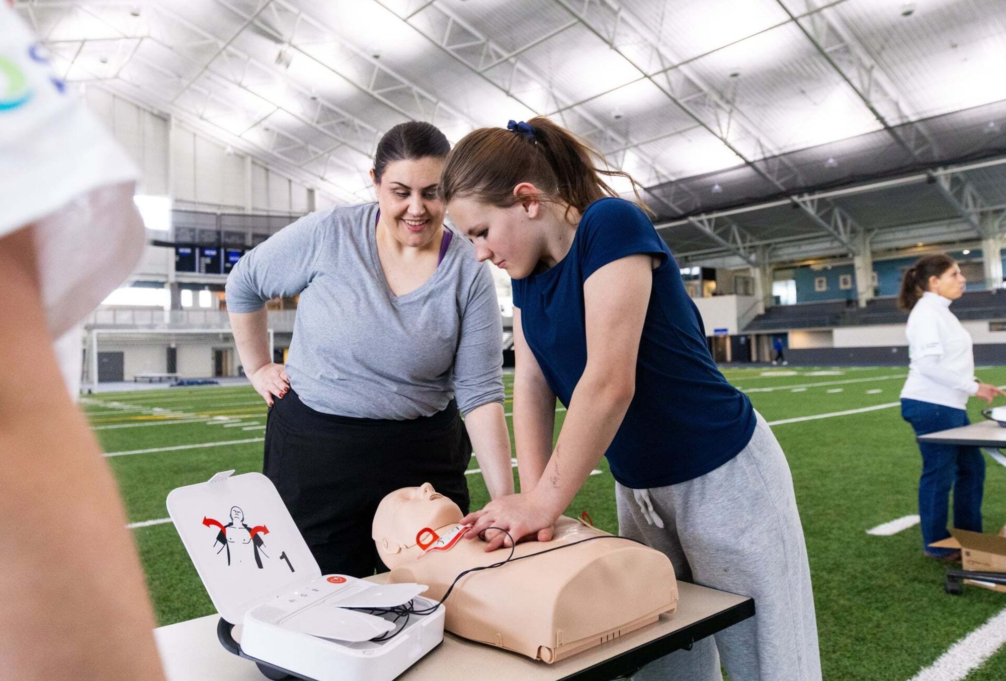 Here are three versions for this photo: Alt Text (for accessibility/SEO): A young woman practices hands-only CPR on a training manikin at an indoor sports facility while a female instructor looks on and smiles during a Champions of the Heart free CPR training event.